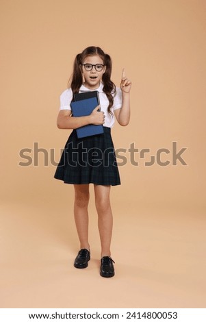 Surprised schoolgirl in glasses with books pointing upwards on beige background