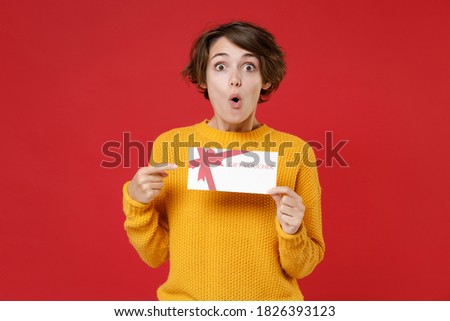 Surprised shocked amazed young brunette woman 20s wearing yellow sweater standing hold in hands pointing index finger on gift certificate isolated on bright red colour background studio portrait