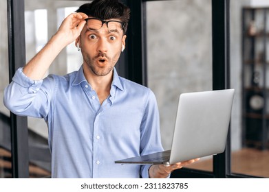 Surprised shocked caucasian businessman, office employee in a shirt, stands in the office, holds an open laptop in his hand, looks in amazement at the camera, raising his glasses, got unexpected news - Shutterstock ID 2311823785