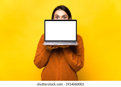 Surprised shocked caucasian young woman, hiding behind blank white screen of laptop, standing on isolated orange background. Copy space.