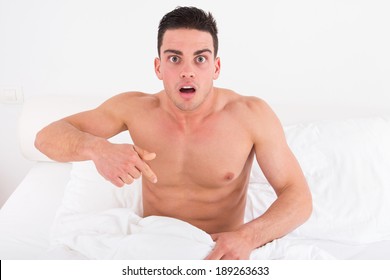 surprised and shocked half naked young man in bed  looking down at his penis under white covers sheet in bedroom. Concept photo of male sexuality and man sex problems, domestic atmosphere.