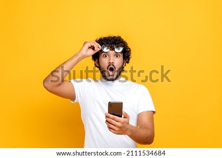 Surprised shocked Indian guy in white t-shirt, holds a smartphone in hand, takes off his glasses and looks in amazement at the camera, received an unexpected message or news,isolated orange background Stock photo © 