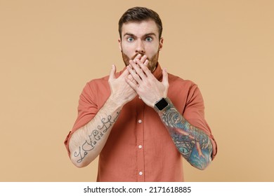 Surprised shocked oops young brunet man 20s he has short haircut earrings wears orange shirt cover mouth with hand isolated on plain pastel light beige background studio portrait. Tattoo translate fun