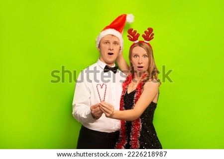 Surprised shocked young people husband and wife or friends wearing Santa Claus hat, reindeer antlers headdress with tinsel, looking at camera and holding Christmas lollipop candy on green background.