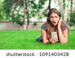 Surprised shocked young woman looking at phone seeing unexpected news or photos with wonder emotion on her face outside in park lying down on green lawn background with copy space. Multicultural model