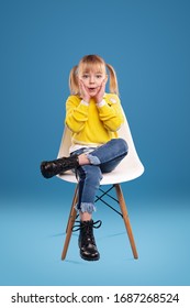 Surprised Small Girl In Bright Yellow Sweater And Jeans And Black Boots Looking At Camera And Touching Cheeks, While Sitting Alone On White Chair Against Blue Background In Studio