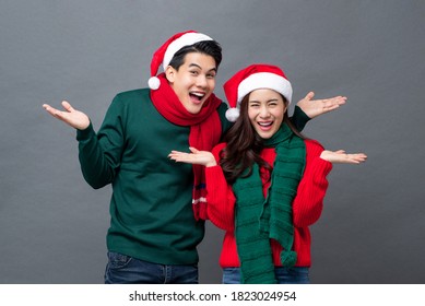 Surprised smiling Asian couple wearing colorful Christmas sweaters and hats doing double open hands gesture in studio isolated gray background - Powered by Shutterstock