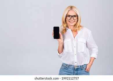 Surprised Smiling Happy Senior Mature Woman In Casual Showing Blank Smartphone Screen While Looking At The Camera Isolated Over White Grey Background. Using Phone.