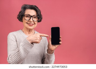 Surprised smiling happy senior mature aged senior woman in casual showing blank smartphone screen while looking at the camera isolated over pink background. Using phone. - Powered by Shutterstock