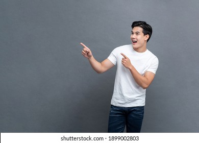 Surprised smiling young handsome Asian man in white t-shirt looking and pointing hands aside to empty space in isolated studio gray background