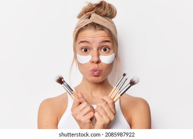 Surprised tender woman keeps lips folded holds cosmetic brushes applies beauty patches under eyes to remove wrinkles stands bare shoulders wears headband casual t shirt isolated over white background. - Powered by Shutterstock