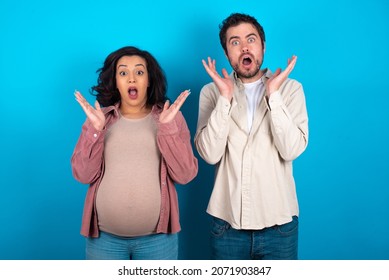 Surprised Terrified Young Couple Expecting A Baby Standing Against Blue Background Gestures With Uncertainty, Stares At Camera, Puzzled As Doesn't Know Answer On Tricky Question, People, Body Language