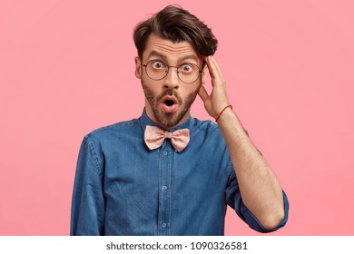 Surprised unshaven hipster male looks in bewilderment, wears glasses and denim shirt, being shocked to see high price, isolated on pink background. Studio shot. Fashionable man in elegant clothes