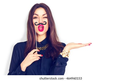 Surprised Winking Model Girl Holding Funny Mustache On Stick And Showing Empty Copy Space On Open Hand Palm For Text, White Background. Girl Presenting Point. Proposing Product. Advertisement Gesture