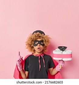 Surprised Woman With Curly Hair Wears Helmet Eye Mask Rubber Gloves And Cloak Pretends Having Superpower And Being Superhero Indicates Above On Blank Empty Space Holds Plunger With Toilet Paper