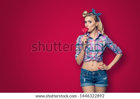 Surprised woman eating heart shape lollipop. Girl in pin up cloth. Blond model at retro fashion and vintage concept. Dark red color background. Copy space for some advertise slogan or text. 