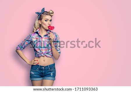 Surprised woman eating heart shape lollipop. Lovely girl in pin up. Blond model at retro fashion and vintage concept. Pink color studio background. Copy space for some ad slogan or text. 