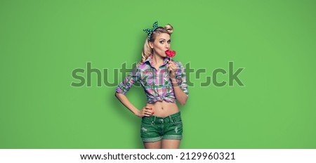 Surprised woman eating heart shape lollipop. Girl in pin up cloth. Blond model at retro fashion and vintage concept. Green color background. Copy space for advertise. 