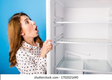 Surprised Woman Looking Into An Empty Fridge On A Blue Background. Quarantine Fasting. Out Of Stock