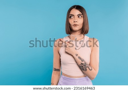 surprised woman showing something and looking away, stylish and tattooed model with nose piercing pointing with finger on blue background, generation z, emotional