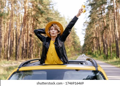 Surprised Woman Standing Out Of Yellow Car Sunroof. Amazed Caucasian Girl Enjoying Road In Woods Front View. Young Blond In Leather Jacket And Hat Express Emotion At Auto Moon Roof