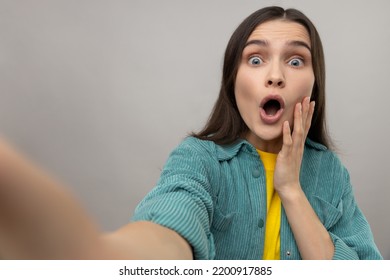 Surprised Woman Taking Selfie With Shocked Facial Expression, Looking With Big Eyes And Open Mouth, POV, Wearing Casual Style Jacket. Indoor Studio Shot Isolated On Gray Background.