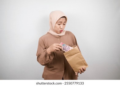 Surprised woman wearing a hijab looking at cash from a paper bag, indicating financial success or a pleasant surprise - Powered by Shutterstock