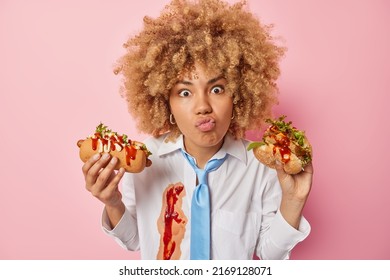 Surprised Wondered Woman Keeps Lips Folded Holds Appetizing Hot Dog And Hamburger Prefers Eating Fast Food Dressed Formally Isolated Over Pink Background. Cheat Meal And Unhealthy Nutrition.