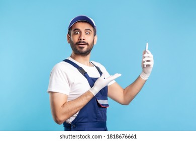 Surprised workman in overalls and gloves pointing mobile phone and looking amazed, shocked by online delivery order app, house repair maintenance services. studio shot isolated on blue background