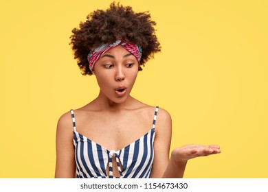 Surprised young African American female with dark curly hair, dressed in fashionable summer clothes, raises palm, pretends holding something, looks with shocked expression, stands in studio.