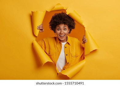 Surprised young African American woman stands in torn paper hole, dressed in stylish clothes, has excited cheerful expression, looks through breakthrough of yellow background. Wow, great news