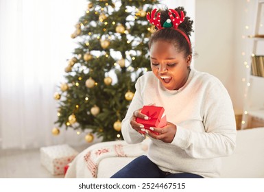 Surprised young African American woman wearing reindeer horns sitting on bed near Christmas tree and opening gift box. Excited black girl holding red present box, celebrating x-mas. Merry Christmas. - Powered by Shutterstock