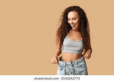 Surprised young African-American woman in loose jeans on beige background. Weight loss concept - Powered by Shutterstock