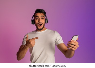 Surprised young Arab guy in headphones pointing at smartphone, shocked over online sale in neon light. Emotional middle Eastern man checking new cool mobile music app