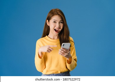 Surprised young Asia lady using mobile phone with positive expression, smiles broadly, dressed in casual clothing and looking at camera on blue background. Happy adorable glad woman rejoices success. - Shutterstock ID 1807522972