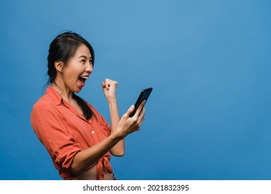 Surprised Young Asia Lady Using Mobile Phone With Positive Expression, Smiles Broadly, Dressed In Casual Clothing And Stand Isolated On Blue Background. Happy Adorable Glad Woman Rejoices Success.