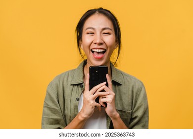 Surprised Young Asia Lady Using Mobile Phone With Positive Expression, Smile Broadly, Dressed In Casual Clothing And Looking At Camera On Yellow Background. Happy Adorable Glad Woman Rejoices Success.