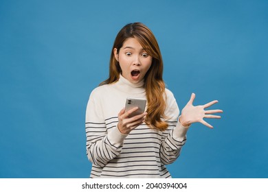 Surprised young Asia lady using mobile phone with positive expression, smiles broadly, dressed in casual clothing and standing isolated on blue background. Happy adorable glad woman rejoices success.