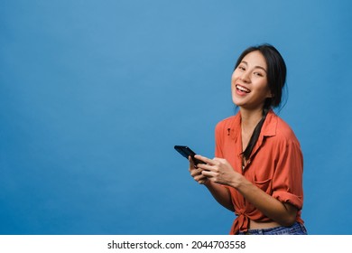 Überraschte junge Asienfrau, die ein Mobiltelefon mit positivem Ausdruck benutzt, im Allgemeinen lächelt, in lockerer Kleidung gekleidet und Kamera auf blauem Hintergrund ansieht. Fröhliche, verehrte Frau freut sich über den Erfolg.