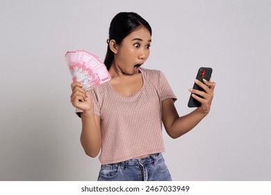 Surprised young Asian woman in casual t-shirt using smartphone, reading promo online and holding in hand fan of cash money in rupiah banknotes isolated on white background. Financial business concept