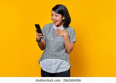 Surprised young Asian woman holding mobile phone with open mouth on yellow background
