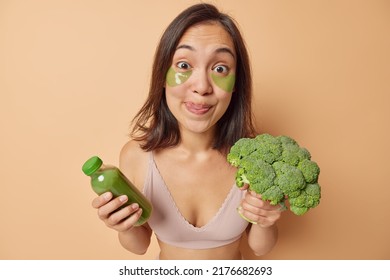 Surprised Young Asian Woman Holds Bottle Of Fresh Detox Beverage Made Of Broccoli Makes Homemade Smoothie Licks Lips Applies Green Hydrogel Patches Under Eyes Isolated Over Beige Background.