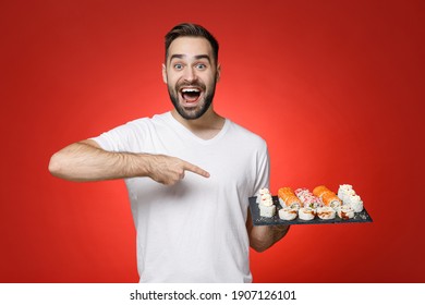 Surprised Young Bearded Man 20s In Casual White T-shirt Point Index Finger Camera On Makizushi Sushi Roll Served On Black Plate Traditional Japanese Food Isolated On Red Background Studio Portrait