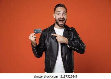 Surprised young bearded man in basic white t-shirt black leather jacket standing pointing index finger on credit bank card looking camera isolated on bright orange colour background studio portrait