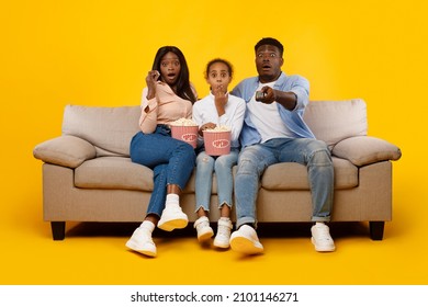 Surprised Young Black Family Of Three People Watching Movie, Excited Parents Sitting With Daughter On Sofa, Eating Popcorn From Bucket Holding Remote Control Isolated On Yellow Orange Studio Wall