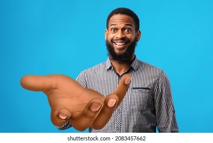 Surprised Young Black Man Standing With Big Outstretched Hand On Blue Studio Background, Mockup For Your Product. Millennial African American Guy Offering Help, Giving Or Asking For Something