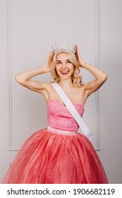 Surprised Young Blonde Woman Wearing A Crown, Pink Dress And A White Ribbon, Smiling. Beauty Contest Winner Concept.