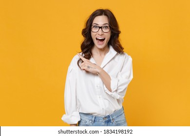 Surprised Young Brunette Business Woman In White Shirt Glasses Isolated On Yellow Background Studio Portrait. Achievement Career Wealth Business Concept. Mock Up Copy Space. Point Index Finger Aside