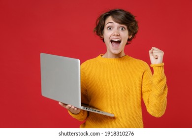 Surprised Young Brunette Woman 20s Wearing Basic Casual Yellow Sweater Standing Working On Laptop Pc Computer Doing Winner Gesture Looking Camera Isolated On Bright Red Background Studio Portrait