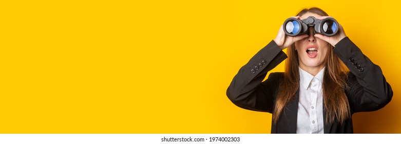 surprised young business woman looking through binoculars on yellow background.